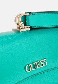 Teal bag with smooth texture, gold hardware, and a logo detail. Short strap with a sleek attachment point, highlighting modern design features.