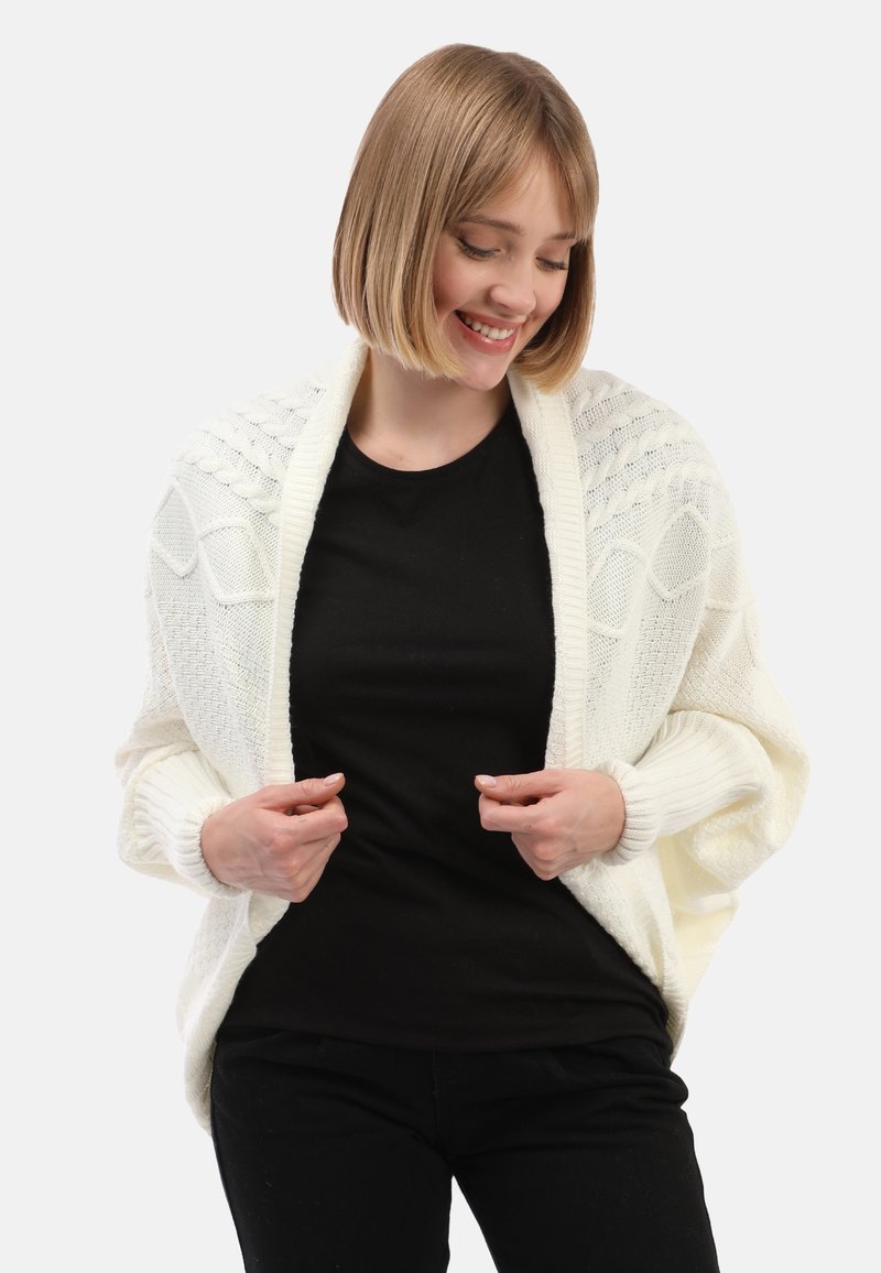 Weißer gestrickter Cardigan mit strukturierten Mustern, offener Vorderseite und gerippten Bündchen. Über einem schwarzen Oberteil getragen, das eine lockere Passform hat.