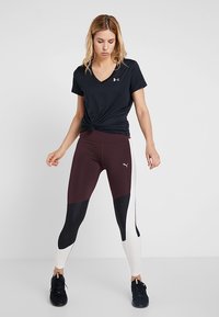 Schwarzes V-Ausschnitt-T-Shirt, am Bund geknotet, kombiniert mit hoch taillierten weinrot-schwarzen Leggings mit weißen Seitenpaneelen und Logo.