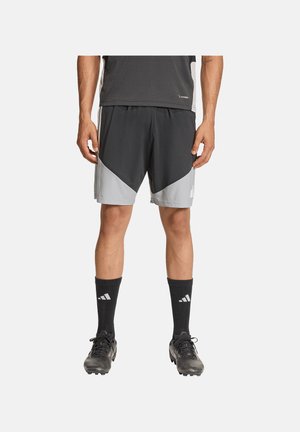 Mann trägt schwarze und graue Sportshorts, schwarze Adidas-Socken und schwarze Stollenschuhe, steht vor einem schlichten Hintergrund.