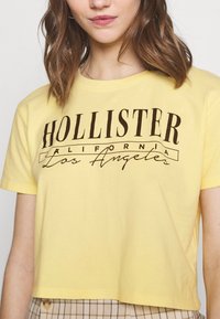 Žluté zkrácené tričko z bavlny s tmavě hnědým potiskem "HOLLISTER" a "California Los Angeles" na přední straně. Krátké rukávy a kulatý výstřih.
