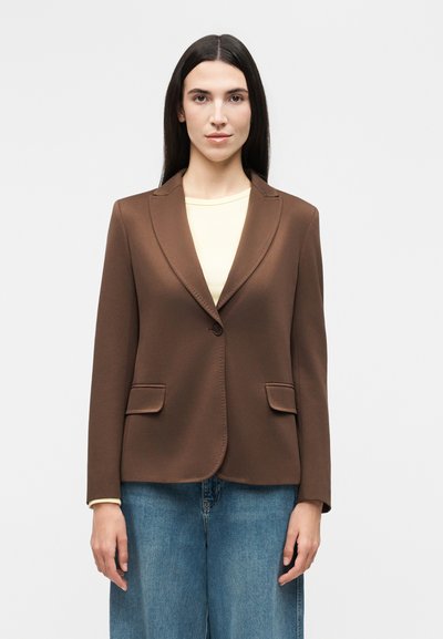 Femme aux longs cheveux foncés portant un blazer marron par-dessus un haut clair et un jean bleu, debout devant un fond blanc uni.