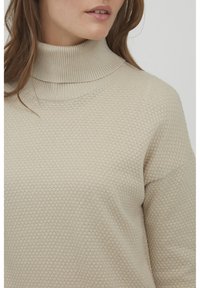 Beige Strickpullover mit hohem geripptem Kragen, der ein strukturiertes Wabenmuster und einen lockeren Schnitt aufweist.