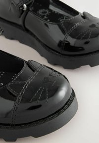 Des chaussures en cuir verni noir avec un bout rond, un design de visage de chat brodé, une semelle en caoutchouc noir texturé contrastant et une sangle ajustable.