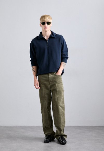 Blond man wearing black sunglasses, navy quarter-zip sweater, olive cargo pants, and black shoes standing against a plain gray background.