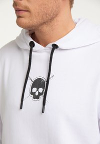 Hvid hættetrøje med kappe, prydet med et sort kraniegrafik med tværsbones, tykke sorte snore og metalspidser. Glat stoftekstur.