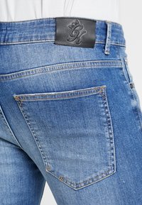 Pantalones vaqueros de mezclilla en azul claro con una superficie texturizada. Presentan un parche de cuero negro con logo en la cinturilla y un diseño en el bolsillo trasero.