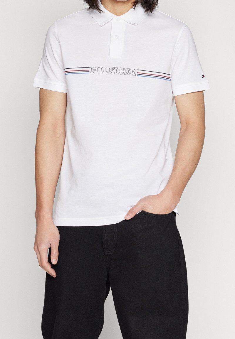 Polo blanc en coton, avec col et manches courtes, ornée d'une bande horizontale bleue et rouge et du texte "HILFIGER" sur la poitrine.