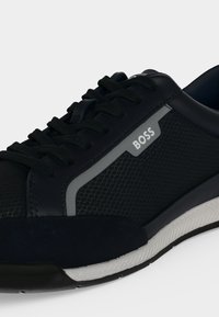 Gros plan sur une basket noire avec des panneaux latéraux texturés, des lacets noirs, une semelle blanche et une petite étiquette grise marquée BOSS.