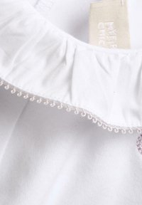 Tessuto bianco con colletto arricciato, caratterizzato da un delicato bordo smerlato. Etichetta visibile con la scritta "MY FIRST CHIC" stampata. La consistenza appare morbida.