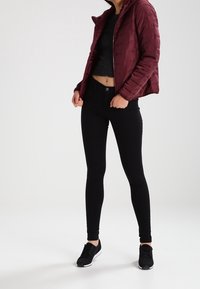 Bordeaux pufferjakke, lynlås lukning, tætsiddende sort top, sorte skinny jeans og sorte træningssko. Glat tekstur, minimalistisk design.
