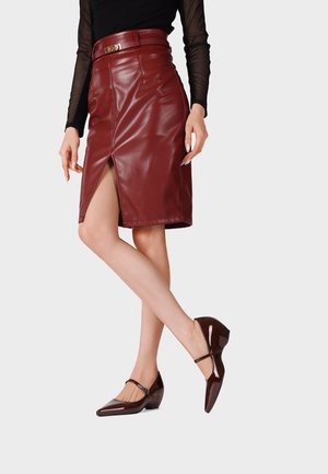 Donna con gonna in pelle marrone a vita alta con spacco frontale, top nero trasparente a maniche lunghe e scarpe Mary Jane marroni verniciate.