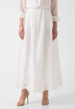 Kobieta ubrana w białą haftowaną długą spódnicę, białą bluzkę z podwiniętymi rękawami, złotą bransoletkę i złote sandały na wysokim obcasie.