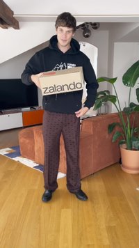Osoba drží béžovú krabicu Zalando, oblečená v čiernej mikine a vzorovaných hnedých nohaviciach. Izba má drevenú podlahu a zelené rastliny.