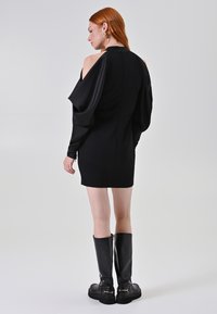 Schwarzes Minikleid mit langen, drapierten Ärmeln und hohem Kragen. Das Model trägt schwarze Stiefel bis über das Knie, die eine glatte Textur und ein elegantes Design zeigen.