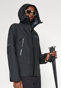 Melnā ūdensnecaurlaidīgā jaka ar kapuci, ar rāvējslēdzēju krūšu kabatu un velcro aprocēm. Modelis tur pārgājienu nūjas, valkājot saulesbrilles.