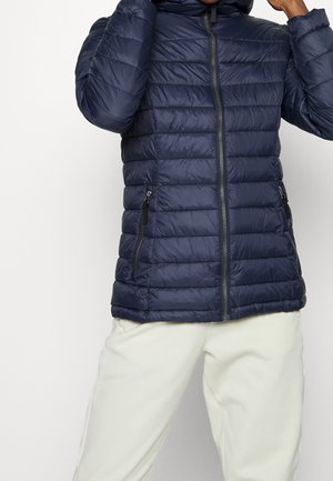 Person wearing a navy blue quilted jacket with front zipper and zippered side pockets, adjusting the hood, paired with white pants.