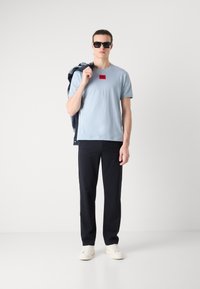 Tricou de culoare albastru deschis, cu mânecă scurtă, având un logo roșu dreptunghiular, asortat cu pantaloni negri și adidași albi. Modelul ține o jachetă din denim peste un umăr.