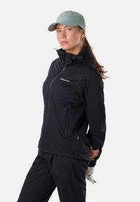 Femme en veste noire à fermeture éclair et pantalon, portant une casquette gris clair, debout les mains dans le dos, légèrement tournée vers la gauche sur fond blanc.