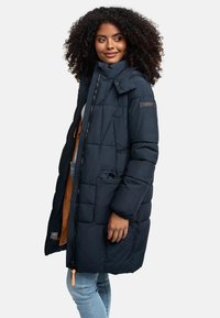 Tumši zila pūkaina jaka ar rāvējslēdzēja aizdari, augstu apkakli un priekšējo kabatu. Raksturīga ir quilted tekstūra un oranža odere.