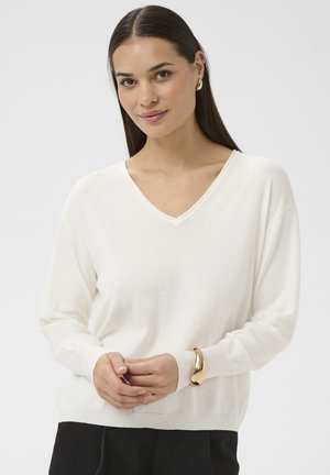 Hosszú sötét hajú fiatal nő fehér V-nyakú pulóvert, fekete nadrágot, arany karkötőt és arany karika fülbevalókat visel, világos háttér előtt állva.