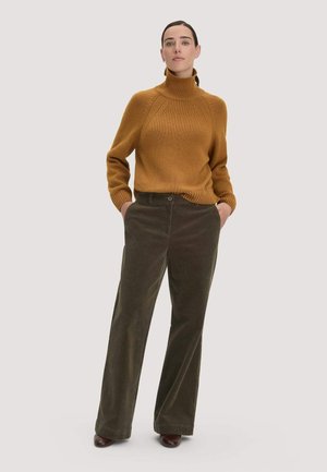 Vrouw staat met handen in de zakken, draagt een mosterdkleurige coltrui en donkergroene wijde corduroy broek met bruine schoenen.