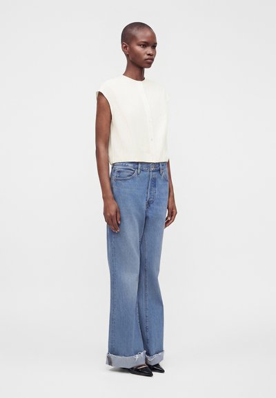 Femme aux cheveux courts portant un chemisier blanc sans manches, un jean bleu large avec revers et des chaussures noires pointues, debout sur un fond uni.