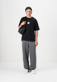 Black T-shirt with a square logo, paired with gray wide-leg sweatpants. Model holds a small black crossbody bag. White shoes complete the look.
