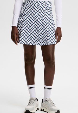 Vrouw draagt een blauw-witte rok met patroon, een wit shirt met lange mouwen, witte sokken met zwarte strepen en witte sportschoenen.