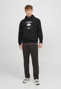Schwarzer Hoodie mit weißem grafischem Druck und Kängurutasche, kombiniert mit dunklen Hosen und mehrfarbigen Turnschuhen, steht vor einem einfarbigen Hintergrund.