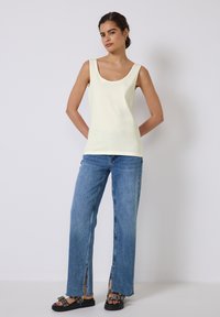 Un débardeur jaune clair sans manches associé à un jean large bleu clair avec des fentes sur les côtés ; le mannequin se tient avec les mains sur les hanches, portant des sandales noires.