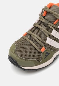 Pánská sportovní obuv s olivově zeleným síťovinovým svrškem, oranžovými akcenty a černou gumovou podrážkou, s bílými pruhy a tkaničkami.