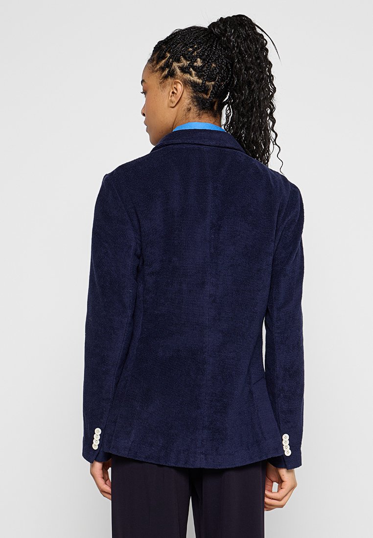 Kobieta z warkoczem w kucyku, ubrana w granatową, teksturowaną marynarkę z białymi guzikami na rękawach, widziana od tyłu na jednolitym tle.