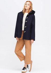Marinblå ullkappa med huva, prydd med guldfärgad hårdvara. Bärs över en vit kabelstickad tröja och kombineras med beige byxor och sneakers.
