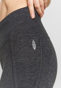 Grå sportleggings i strukturerad tyg. Har en liten vit logo och platta sömmar. Nära vy av midjebandet och benområdet.