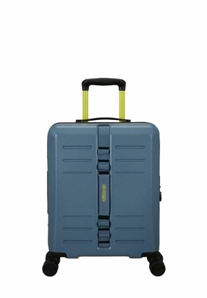 Hard-shell suitcase in dark blue with vertical grooves, yellow retractable handle, and four wheels. Features a travel strap with logo detail.