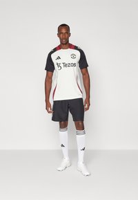 Witte en zwarte voetbalshirt met rode accenten, uitgerust met zwarte shorts, witte sokken met zwarte strepen en witte voetbalschoenen.
