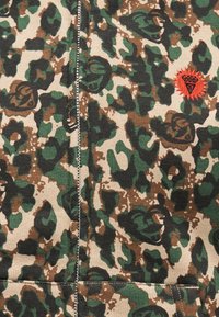 Καμουφλαρισμένο ύφασμα με φερμουάρ, με πράσινες, καφέ και μπεζ κηλίδες, και πορτοκαλί κεντημένο έμβλημα στη δεξιά πλευρά.