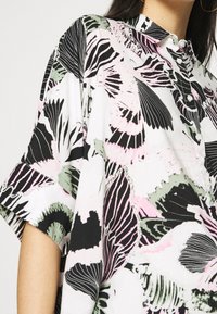 Chemise boutonnée avec un motif floral en noir, rose et vert, manches courtes et col structuré.