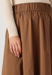 De hand van een vrouw rust op een bruine gestikte rok, terwijl ze een beige geribbelde top met lange mouwen draagt.