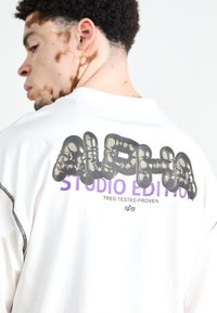 Valkoinen lyhythihainen paita, jossa on suuri musta pörrölogo "ALPHA" ja violetti teksti "STUDIO EDITION" selässä, sekä kontrastivärinen ompelu.}}
