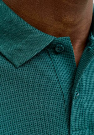 Zeegroen getextureerde poloshirtkraag en knopenlijst op een donkere huid, met zichtbaar detail van de stofbreiwerk en twee zichtbare knopen.