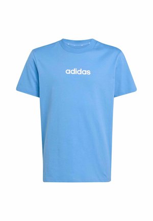 Adolescente de pie con una mano en el bolsillo, vistiendo una camiseta azul de Adidas y pantalones deportivos gris claro sobre un fondo liso.