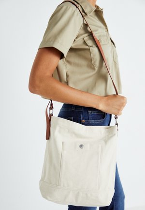 Personne portant une chemise kaki et un jean, portant un sac bandoulière en toile blanc cassé avec une lanière brune et une poche avant, sur un fond uni.