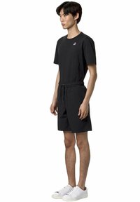 T-shirt nera con un piccolo logo sul petto, abbinata a pantaloni corti neri con vita elastica e tasche laterali, indossati con sneakers bianche.