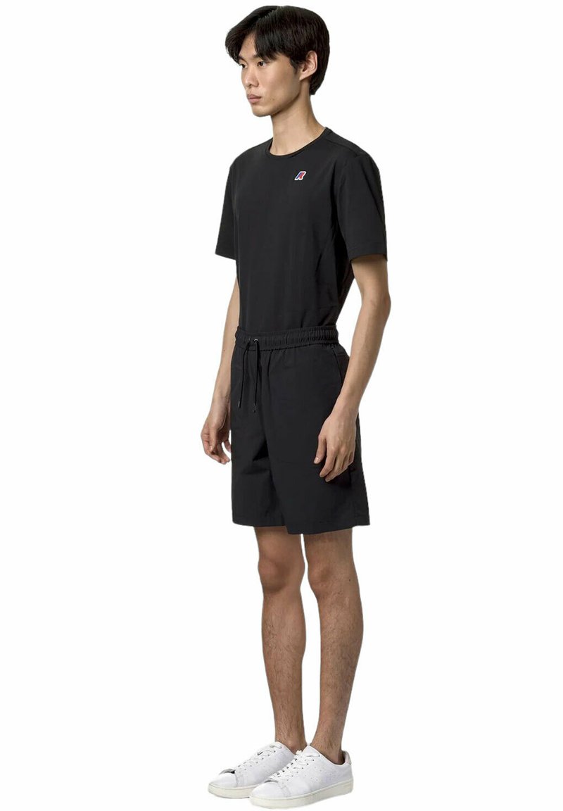 T-shirt nera con un piccolo logo sul petto, abbinata a pantaloni corti neri con vita elastica e tasche laterali, indossati con sneakers bianche.