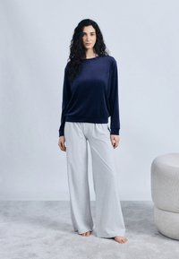 Γυναίκα με μακριά, κατσαρά μαλλιά που φοράει μπλε navy μπλούζα με μακριά μανίκια και φαρδιά ανοιχτόγρια παντελόνια, όρθια ξυπόλυτη σε χαλί κοντά σε στρογγυλή καρέκλα.