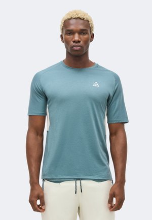 Mann med lyst blondt krøllete hår iført en turkis kortermet treningsskjorte og lyse beige shorts, stående foran en ensfarget bakgrunn.