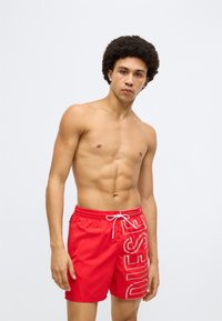 Punased ujumispüksid läikiva viimistlusega, millel on valge logotrükk küljel, elastne vöökoht paelaga ja mugav lõige.