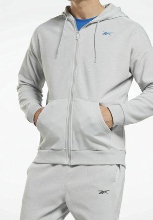 Homme portant un sweat à capuche zippé gris clair et un pantalon de jogging assorti avec des logos de la marque sur la poitrine et la cuisse, mains dans les poches du sweat.